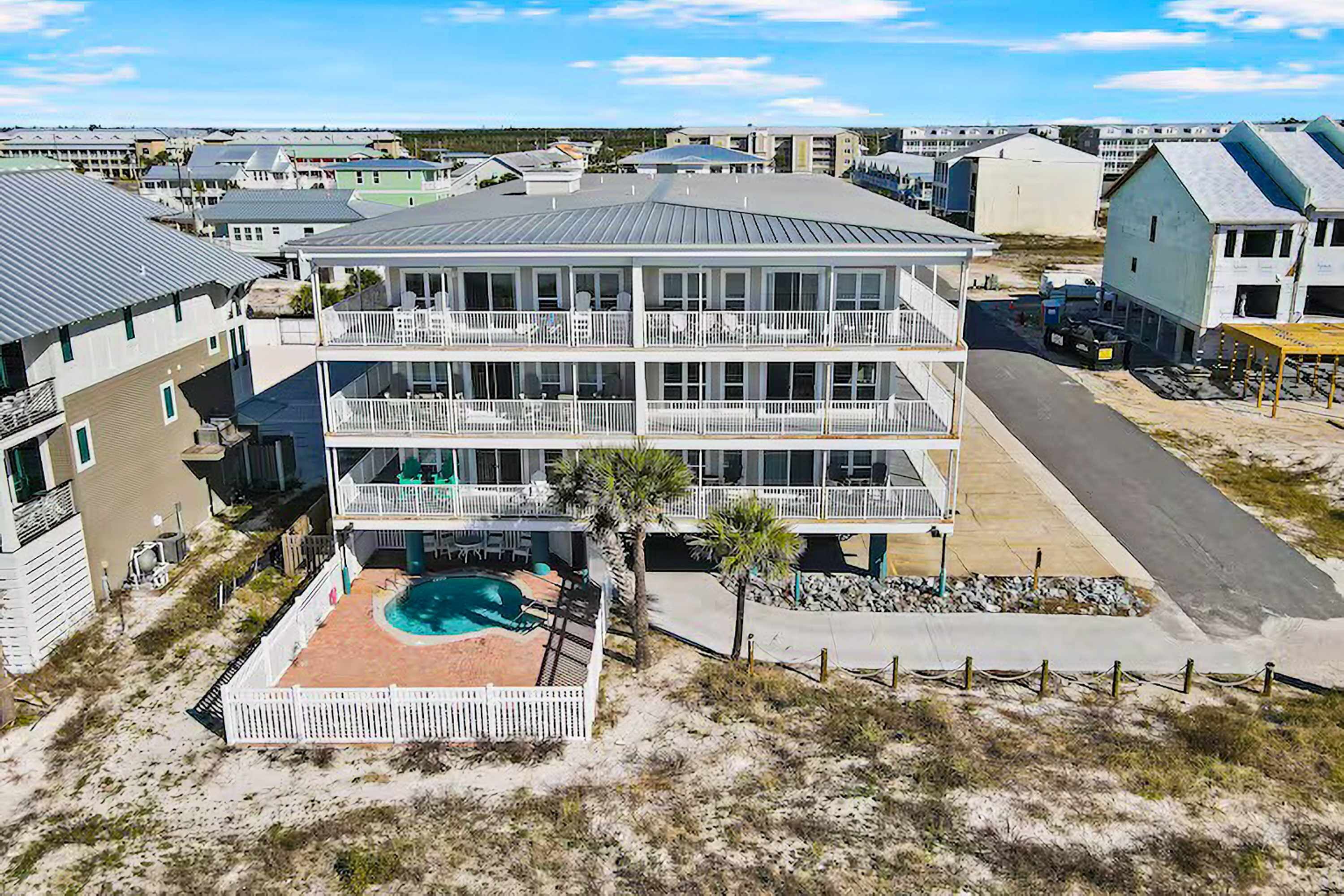 A multi-story building with a pool, surrounded by a sandy area and other residential buildings.