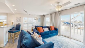 A modern living room with a large blue sofa set, a dining area with bar stools, and a view of the city through large windows.