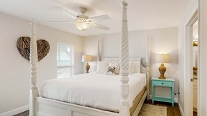 A four-poster bed with a ceiling fan, a small side table with a lamp, and a heart-shaped wall decoration.