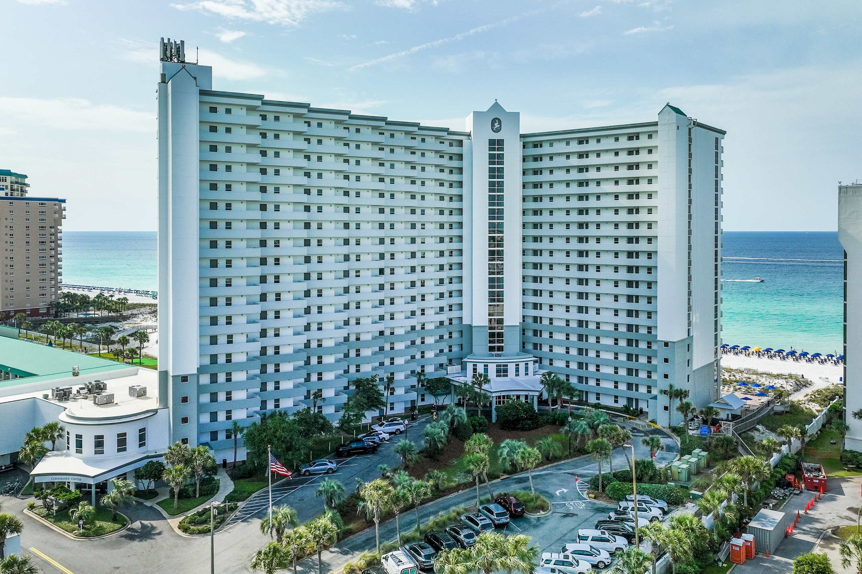 A large hotel with a central tower, multiple floors, and a parking area with cars.