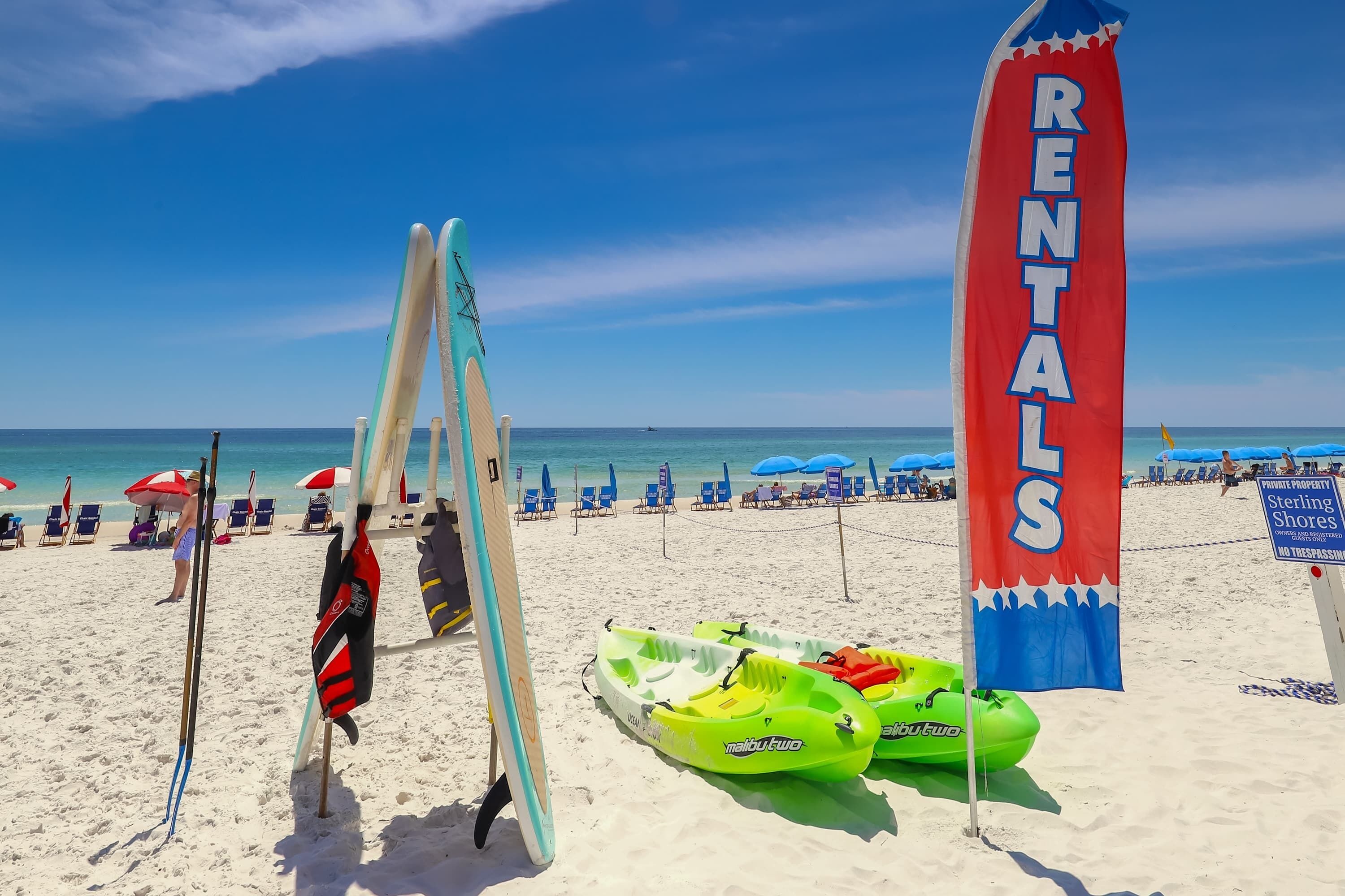A beach with rental surfboards and kayaks, a 'RENTALS' sign, and beach chairs under umbrellas.