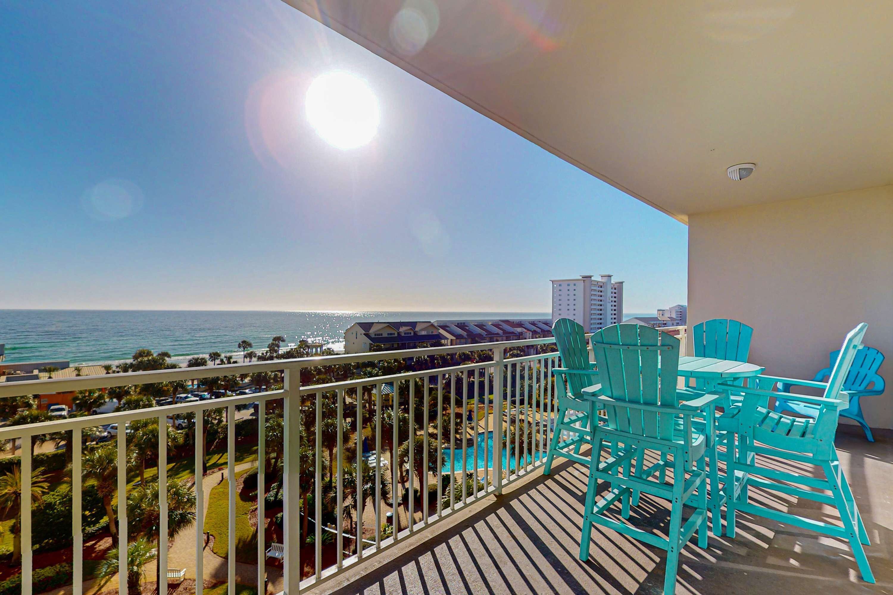 A balcony with turquoise chairs and a table overlooking a beach and ocean.