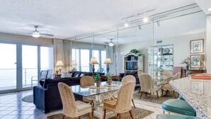 Een ruime woonkamer met een glazen eettafel, omringd door rieten stoelen, een donkere bank en een televisie.