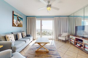 A living room with a sofa, a coffee table, a TV, and a balcony with curtains. - Gulf-view Sterling Shores condo with pool, gym & beach access (Destin)