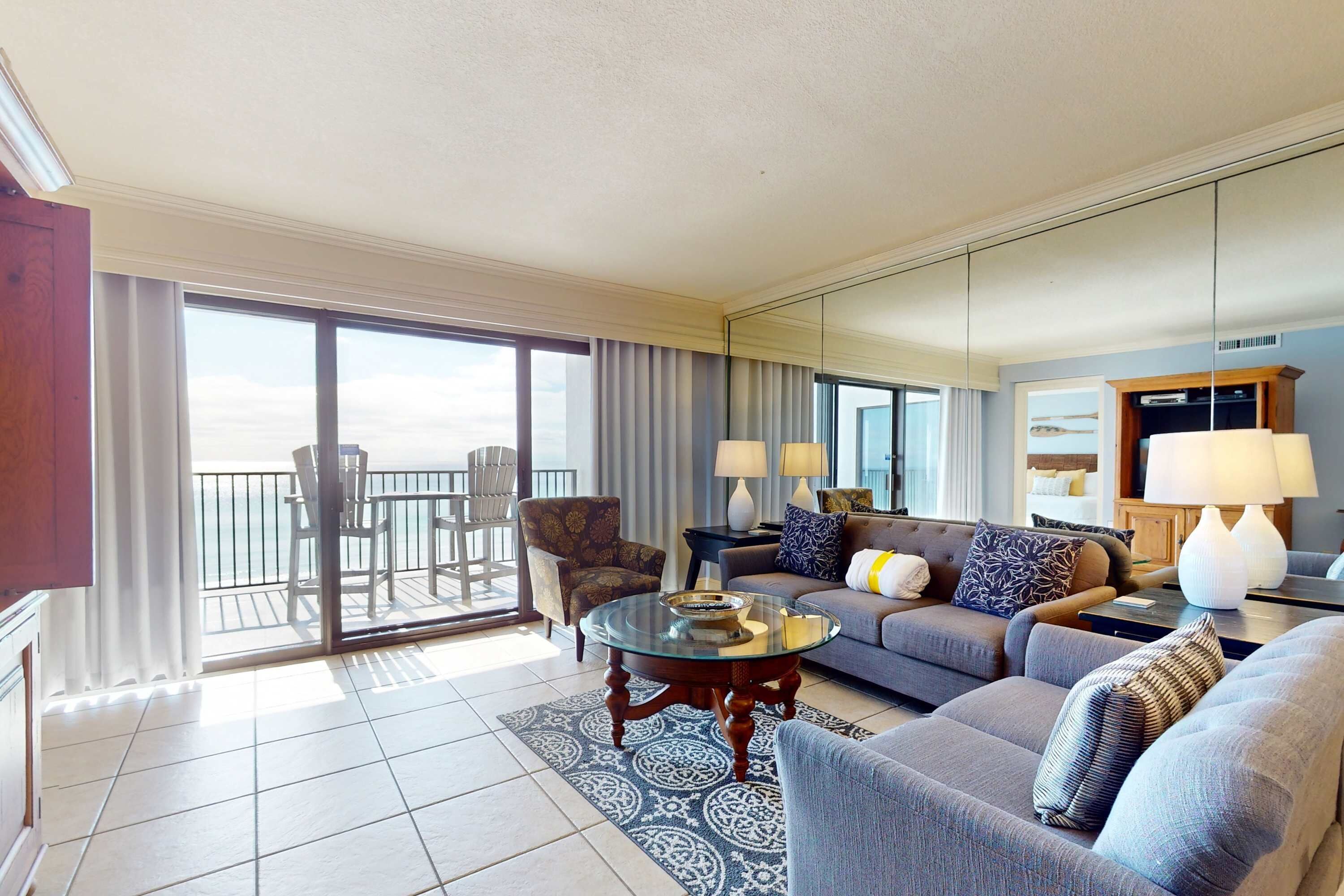 A hotel living room with a sofa, armchairs, a glass coffee table, and a view of the balcony.