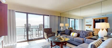 A hotel living room with a sofa, armchairs, a glass coffee table, and a view of the balcony.
