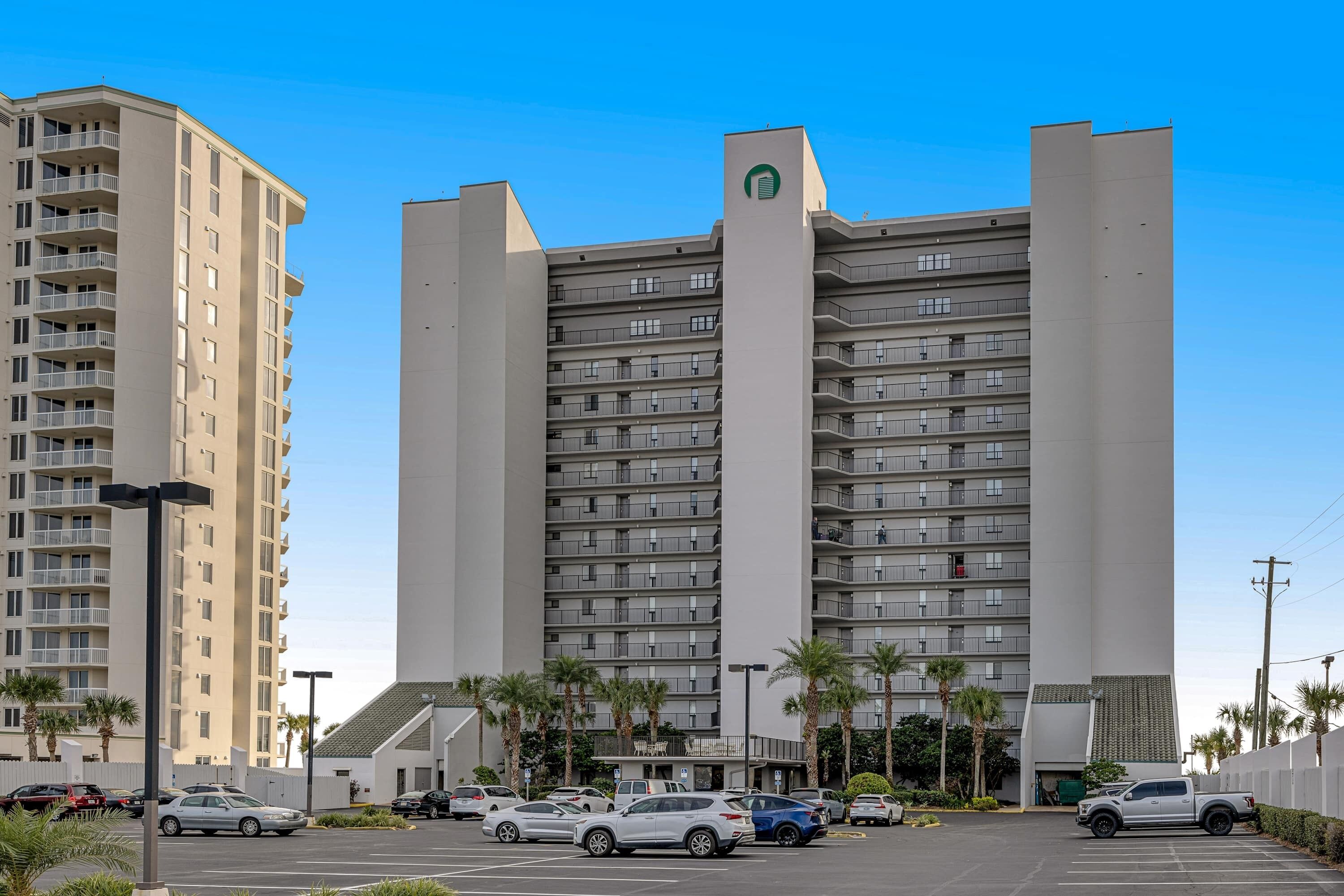 Ein mehrstöckiges Hotel mit Parkplatz vor der Fassade, einige Autos und Palmen sind zu sehen.