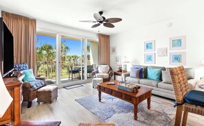 A spacious living room with a balcony, a ceiling fan, and a view of palm trees. - Marvelous Gulf-front condo with amazing views, pool, snack bar, & fitness room (Destin)