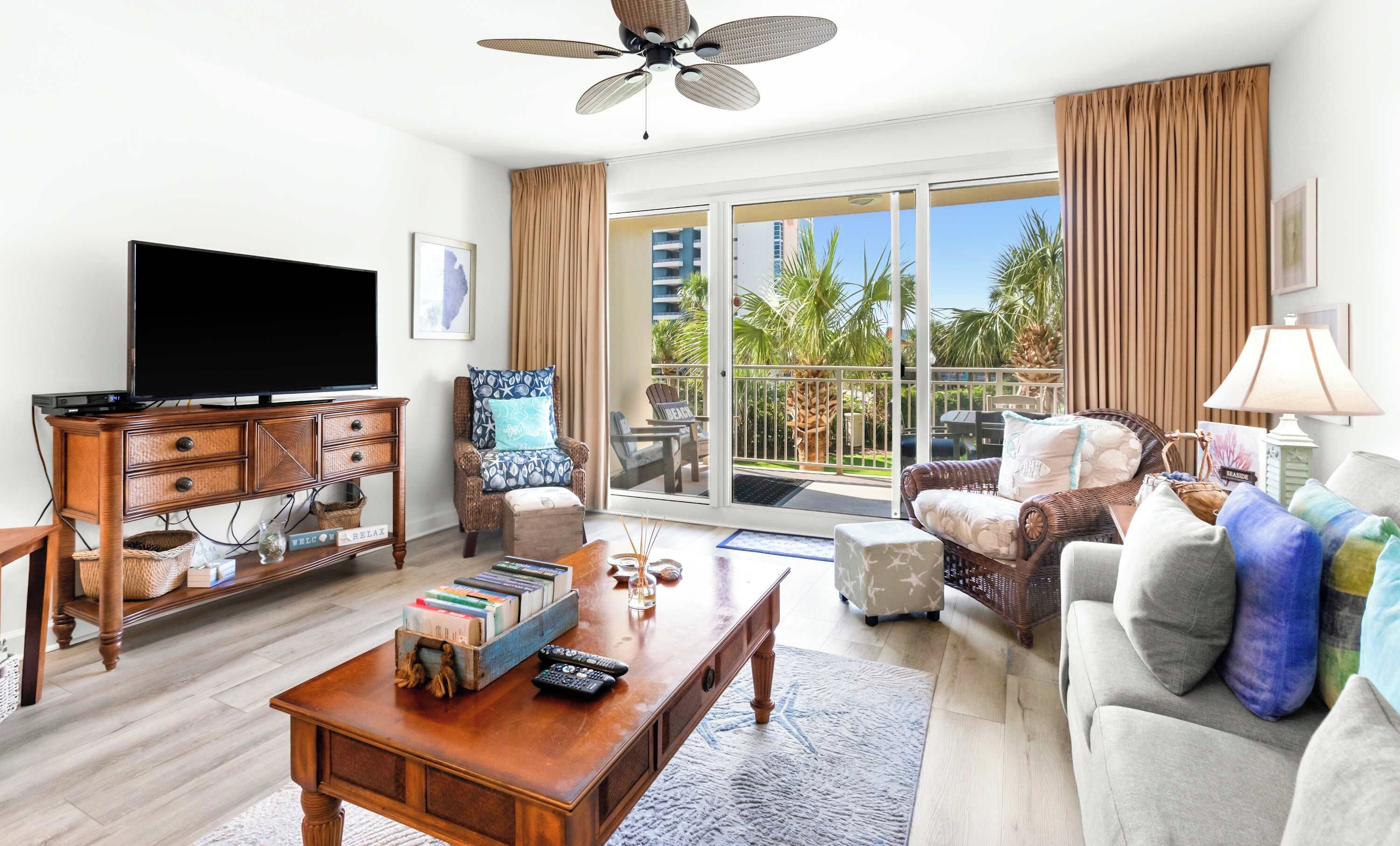 A spacious living room with a flat-screen TV, wooden furniture, a balcony with a view of palm trees, and a ceiling fan.