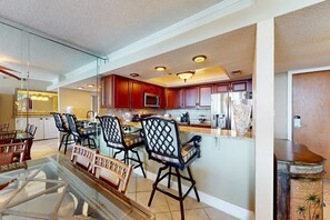 A modern kitchen with wooden cabinets, a central island, and a dining area with chairs and a glass table. - Ocean-view 2nd-floor condo with outdoor pool, gym, hot tub, balcony, WD, AC (Destin)