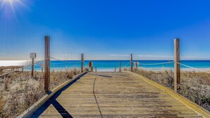Ein hölzerner Steg führt zu einem Strand mit klarem, blauem Wasser und strahlender Sonne.
