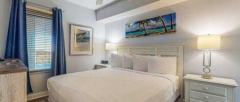 A neatly arranged bedroom with a large bed, a ceiling fan, a nightstand with a lamp, and a painting on the wall.