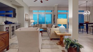 A spacious living room with a large flat-screen TV, comfortable seating, a ceiling fan, and large windows offering a view of the outdoors. - Spacious penthouse condo and private balcony, shared pool&fitness, beach access (Destin)
