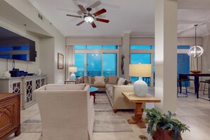 A spacious living room with a large flat-screen TV, comfortable seating, a ceiling fan, and large windows offering a view of the outdoors. - Spacious penthouse condo and private balcony, shared pool&fitness, beach access (Destin)