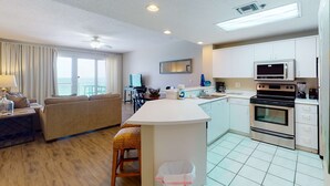 A modern kitchen with a central island, a dining area with a view, and a living room with a sofa and a TV. - Gorgeous beach view home with balcony, pool & fitness room (Destin)