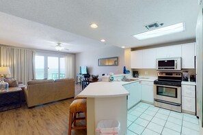 A modern kitchen with a central island, a dining area with a view, and a living room with a sofa and a TV. - Gorgeous beach view home with balcony, pool & fitness room (Destin)