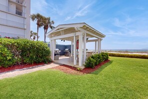 A covered outdoor seating area with a grill, surrounded by well-maintained grass and hedges, near a building with balconies. - Gorgeous beach view home with balcony, pool & fitness room (Destin)
