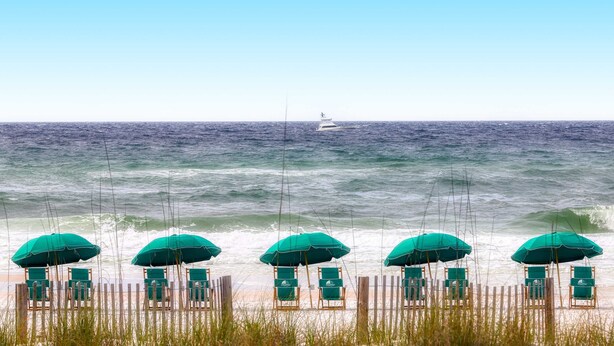 A beach with rows of green umbrellas and beach chairs, a wooden fence, and a boat in the distance. - Gorgeous beach view home with balcony, pool & fitness room (Destin)