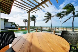 A wooden table and chairs in a covered outdoor area with a view of the ocean and palm trees. - The Point Bungalow on the Sand in Fortim By Carpediem (Fortim)
