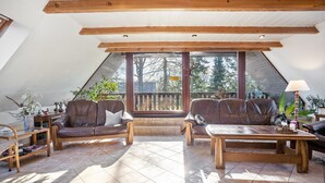 Una habitación acogedora con techo inclinado, vigas de madera, un ventanal grande y una zona de estar con sillones de cuero y una mesa de madera.