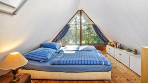 Un dormitorio con techo inclinado, una cama con ropa de cama azul, una mesita de noche con una lámpara, una ventana con cortinas y un pequeño armario con objetos decorativos.