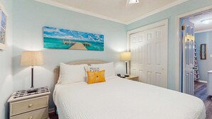 Un dormitorio con una cama, dos mesitas de noche con lámparas, un cuadro de un muelle y una puerta a otra habitación.