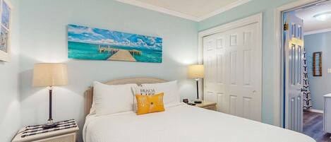 A bedroom with a bed, two bedside tables with lamps, a painting of a pier, and a door to another room.