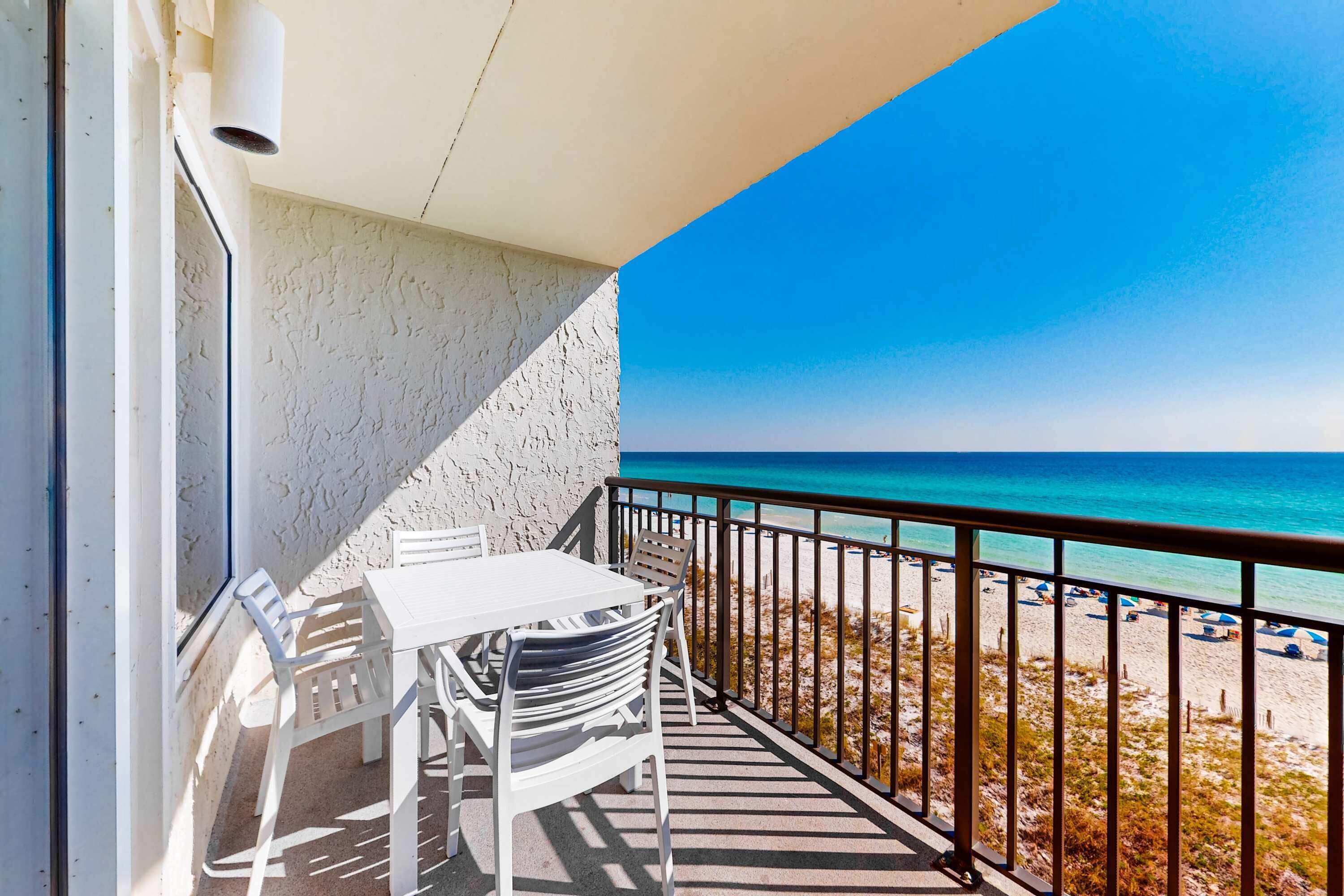 Ein Balkon mit weißem Tisch und Stühlen, der auf einen Strand mit blauem Himmel und türkisfarbenem Meer blickt.