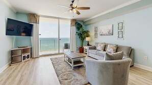 A living room with a TV, a ceiling fan, a sofa, a coffee table, and a view of the ocean through large windows. - Deluxe, beachfront escape with furnished balcony, pools, hot tub & gym (Panama City Beach)