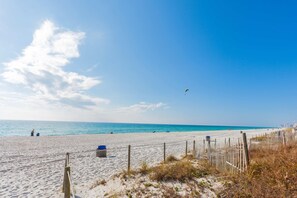 Ein Strand mit blauem Himmel, sandigem Ufer und einer Person, die eine Drachen steigen lässt.