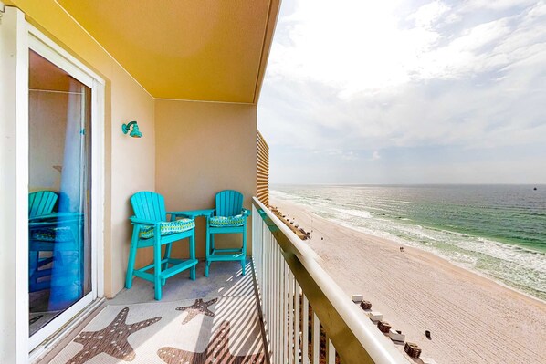 A balcony with turquoise chairs and a starfish-patterned mat, overlooking a beach with the ocean and a clear sky. - Colorful, waterfront condo with pools, gym, & beach access (Panama City Beach)