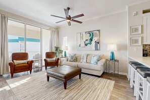 A spacious living room with a large window, a ceiling fan, a sofa, a coffee table, and a bar area with stools. - Luxe Beachfront Resort Condo with Private Beach Access & Top Amenities (Santa Rosa Beach)
