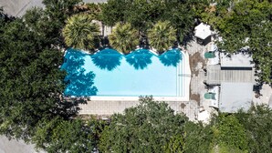 Vista aérea de una piscina rectangular rodeada de abundante vegetación, con un sendero pavimentado y un edificio pequeño a la derecha.