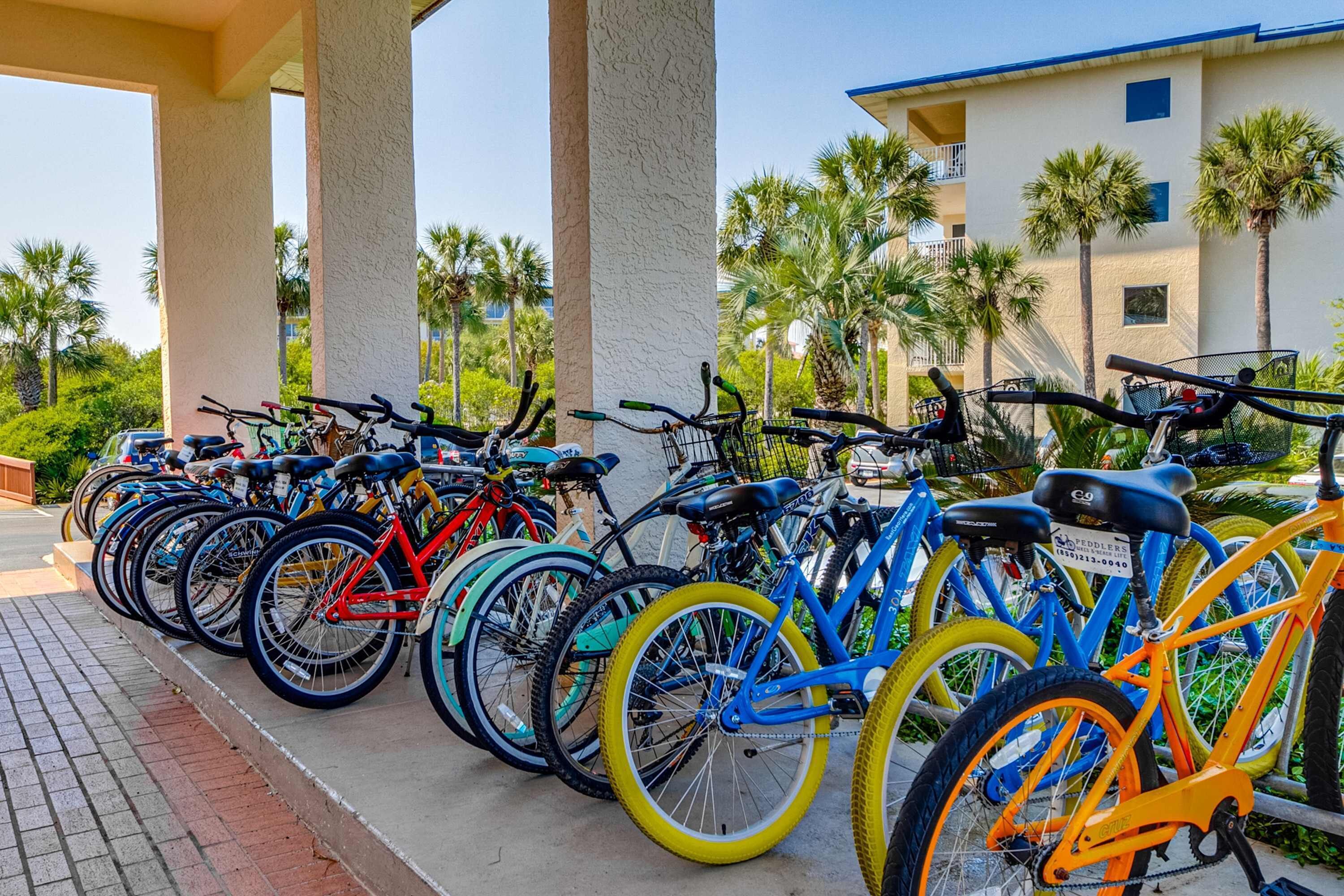 Una hilera de bicicletas estacionadas bajo una zona techada, con palmeras y un edificio al fondo.
