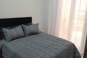 A neatly made bed with a geometric patterned bedspread and pillows, a dark headboard, and sheer curtains allowing natural light. - Honeycomb apartment (Béjaïa)