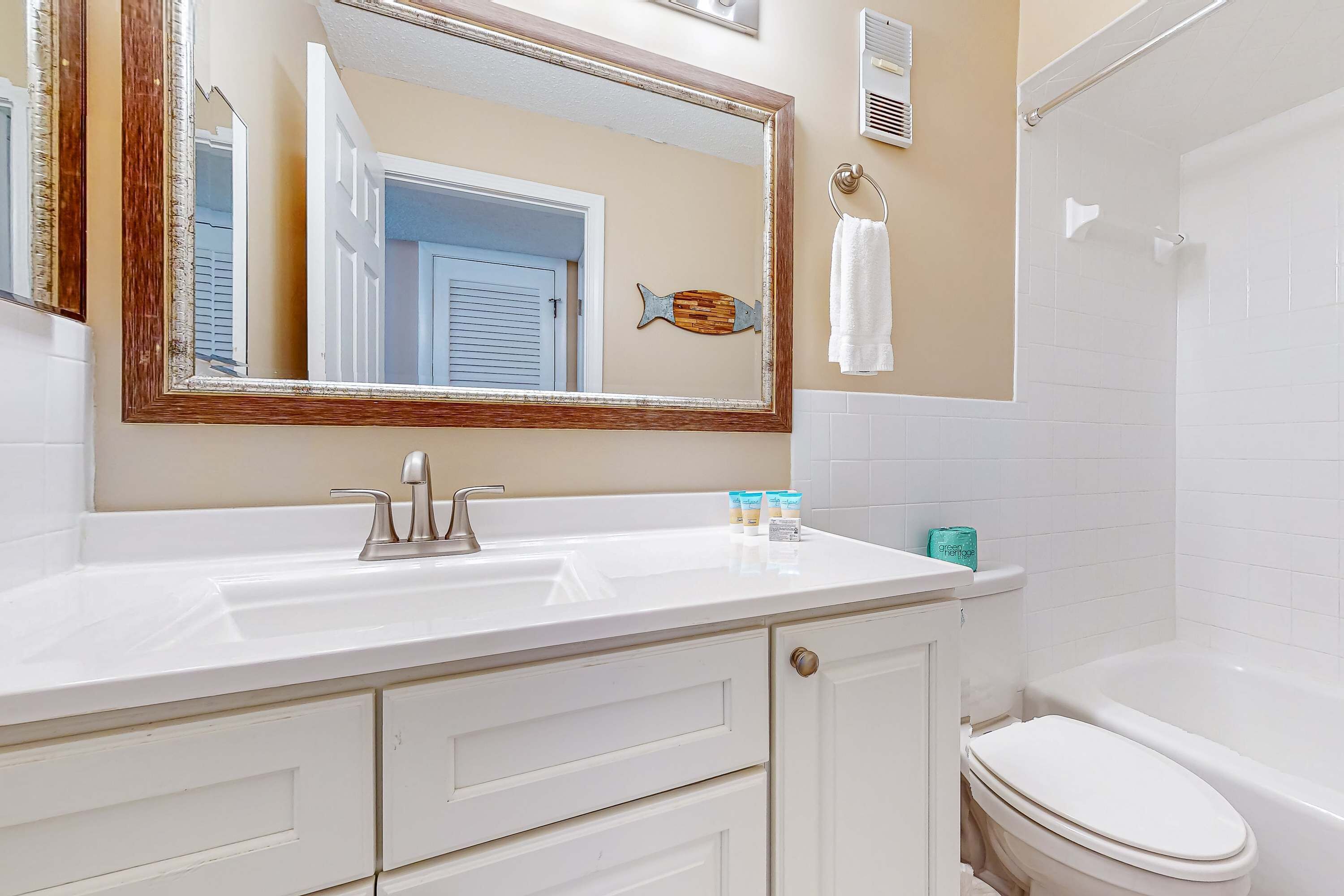 Une salle de bain avec un lavabo blanc, un miroir et des toilettes. On y trouve un porte-serviettes avec une serviette blanche, ainsi qu’un élément décoratif en forme de poisson accroché au mur.