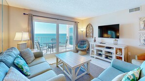 Ein geräumiges Wohnzimmer mit einer Couch, einem Couchtisch und einem Fernseher auf einem weißen Schrank. Durch die Glastüren ist ein Balkon mit Blick sichtbar.