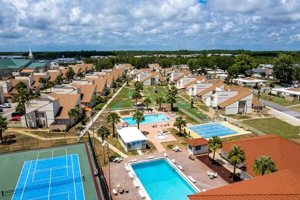 A residential area with multiple houses, a swimming pool, tennis courts, and a church steeple in the background. - Resort condo with putt-putt, gym, pools, hot tub, tennis, & beach access (Panama City Beach)