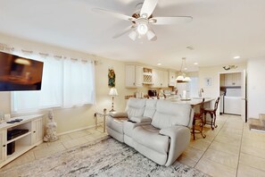 Ein Wohnzimmer mit einer Couch, Fernseher und Deckenventilator. Im Hintergrund ist eine Küche mit Schränken und einem Essbereich zu sehen.
