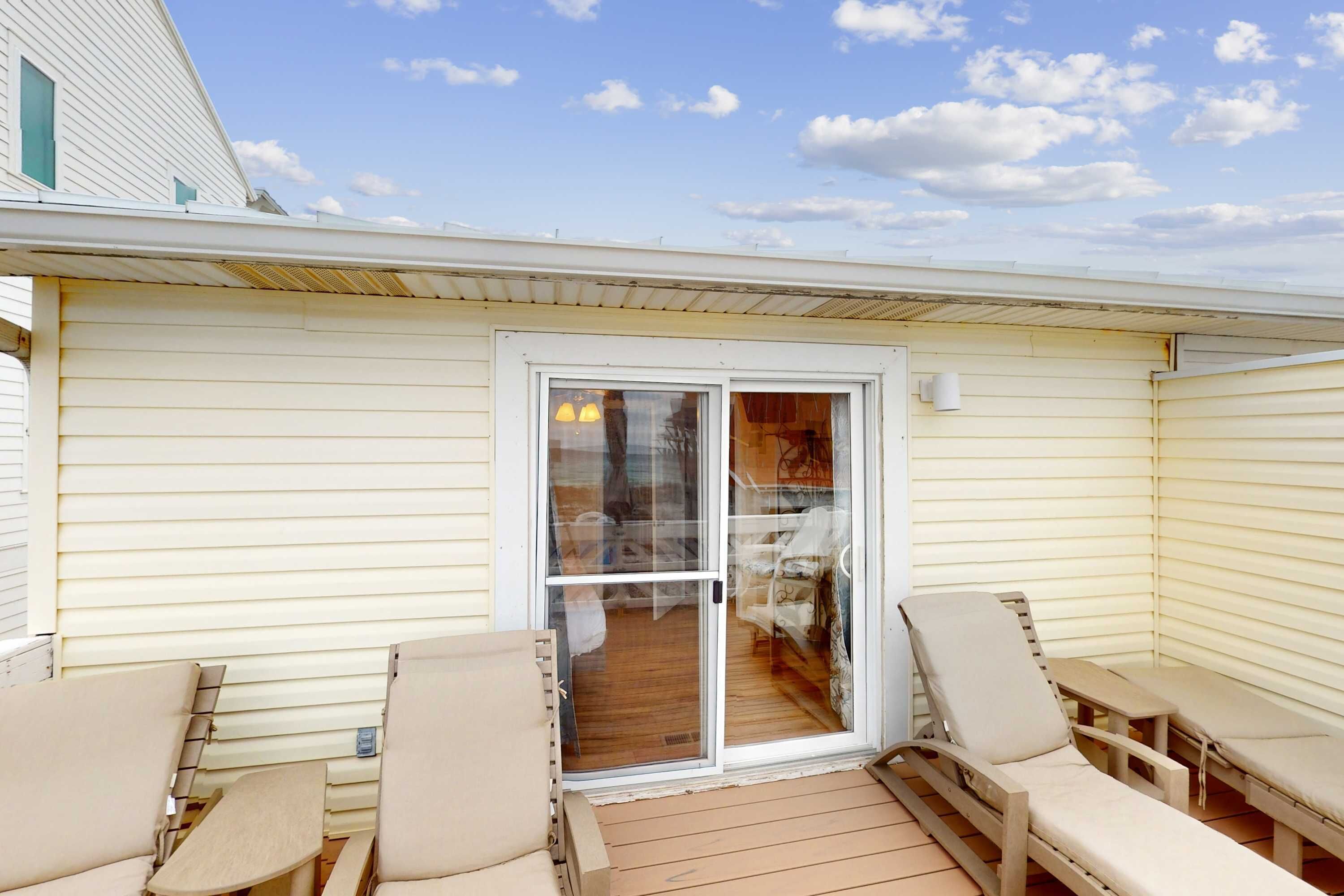 A covered patio with two lounge chairs and a table, a sliding glass door leading to an interior room, and a white exterior wall.