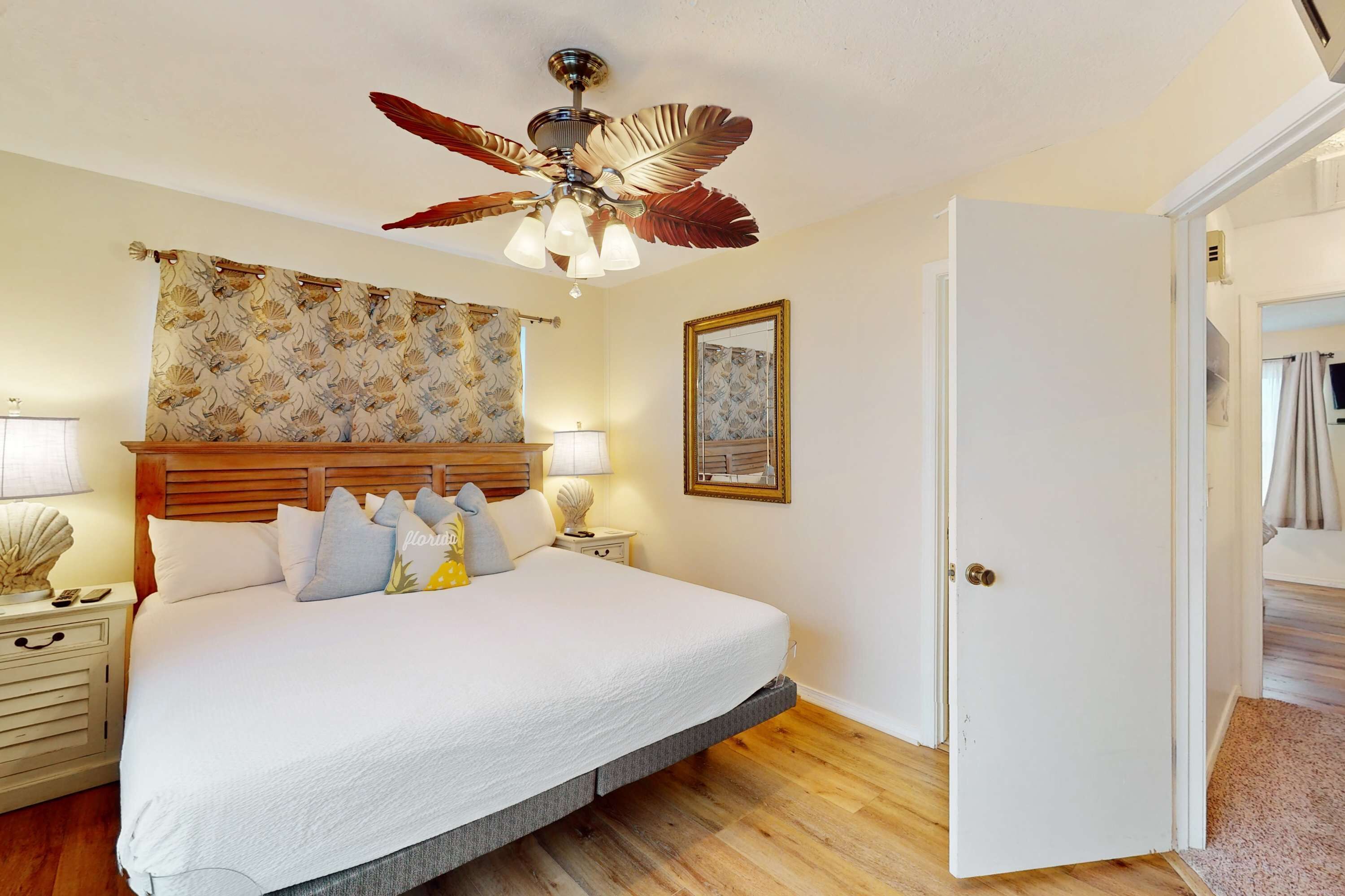 Un dormitorio con una cama grande, un ventilador de techo, una mesita de noche con lámpara y una puerta que da a otra habitación.