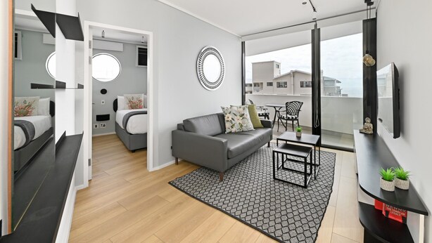 A modern apartment with a grey sofa, a round mirror, and a dining area with a table and chairs. - The Flamingo | Sea Point Prom | One Bed | Balcony (Cape Town)