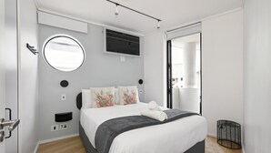 A modern bedroom with a bed, a round mirror, a built-in air conditioning unit, and a small black side table. - The Flamingo | Sea Point Prom | One Bed | Balcony (Cape Town)