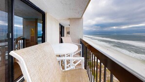Un balcón con una mesa y sillas blancas, con vista a una playa y olas.