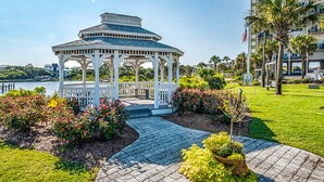 Um gazebo com estrutura branca e teto azul, cercado por jardins bem cuidados e um calçada pavimentada.