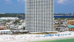 Een hoog, meerlaags hotel met een witte gevel en balkons, gelegen in de buurt van een strand met mensen en parasols.