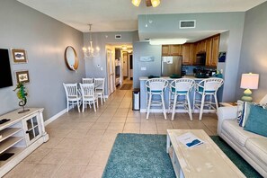 A modern living room with a dining area, kitchen, and a TV mounted on the wall. - Gorgeous beachfront condo, shared pool and beach access (Panama City Beach)