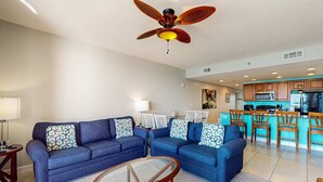 Ein Wohnzimmer mit blauem Sofagarnitur, einem Glastisch und einem Deckenventilator.