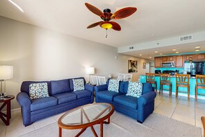 A living room with a blue sofa set, a glass coffee table, and a ceiling fan. - Lovely beachfront resort condo with waterpark, pools, hot tub & beach access (Panama City Beach)