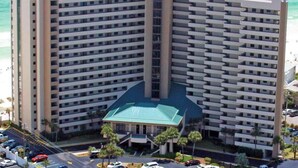 A high-rise hotel with multiple floors, a central entrance, and a parking area with several cars. - 16th floor condo with shared on-site pools, hot tub, restaurant, and bar (Destin)